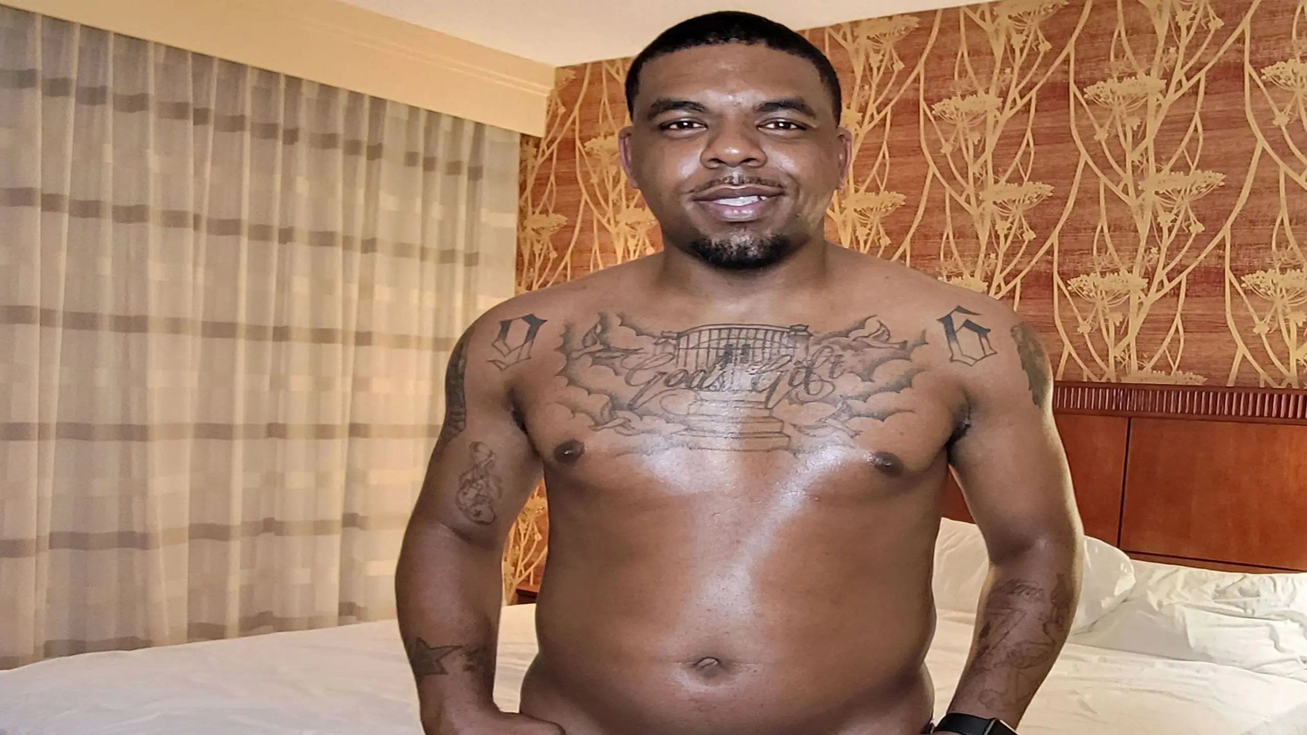 A tattooed man stands confidently in front of a neatly made bed, showcasing his unique body art.