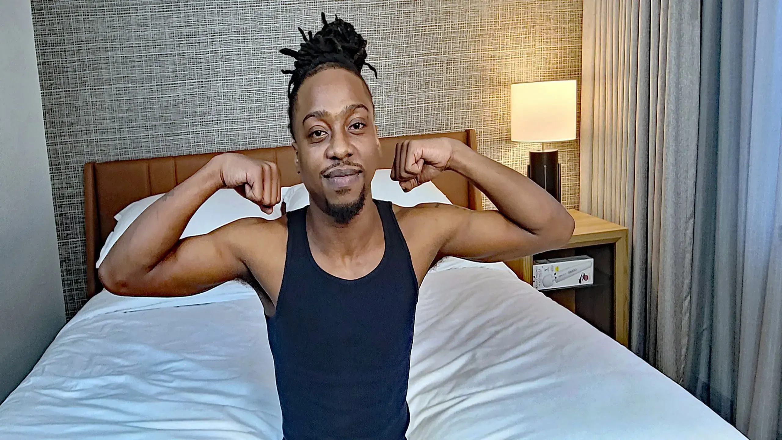 A man strikes a pose in front of a cozy bed, looking relaxed and cheerful.